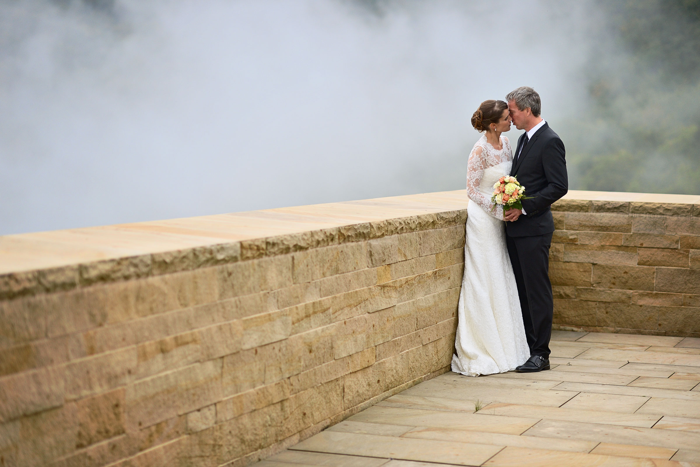 Onelovephoto Hochzeitsfotografie aus Mannheim. Beschreibung: Ein Brautpaar küsst sich an einer Steinmauer vor einem Wasserfall. Lebendige Hochzeitsreportagen für jedes Budget.